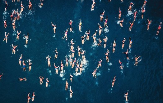 Tientallen mensen liggen zwemmend in diep-blauw water. Waar zwemmen ze heen? De meeste zwemmen van onder in het beeld, naar boven in het beeld. Zwemmen ze echt samen? Wat zit er onder het water?